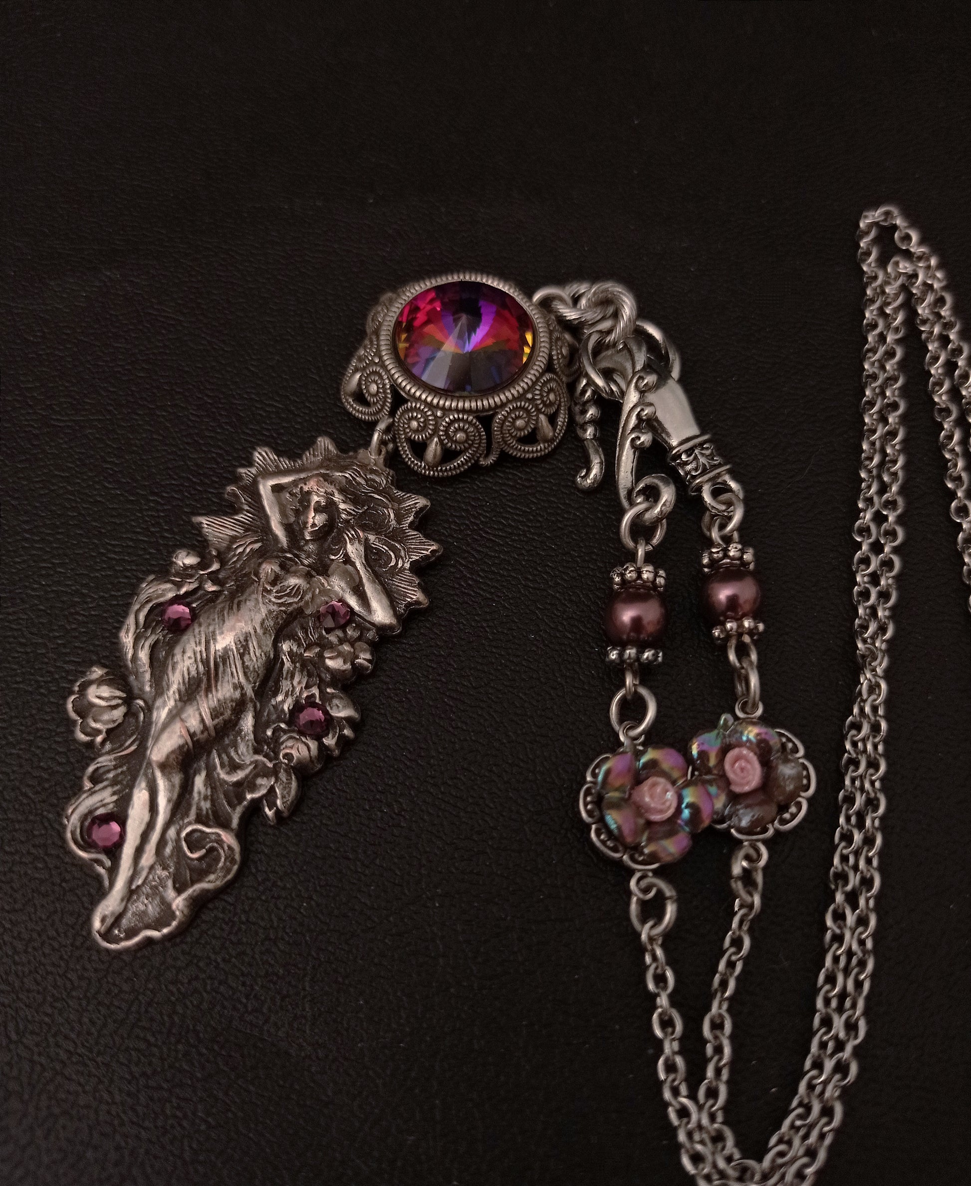 A silver pendant necklace with a detailed woman in flowing robes, set with small purple stones and a large iridescent purple gem. The chain clasps in the front and is further decorated with two lilac pearls and pink roses.