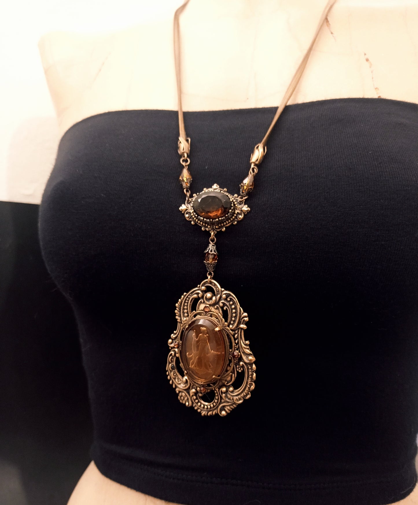 An ornate vintage necklace with a large amber cameo in an intricate brass filigree setting, connected by a smaller amber crystal medallion to gold snake chain.