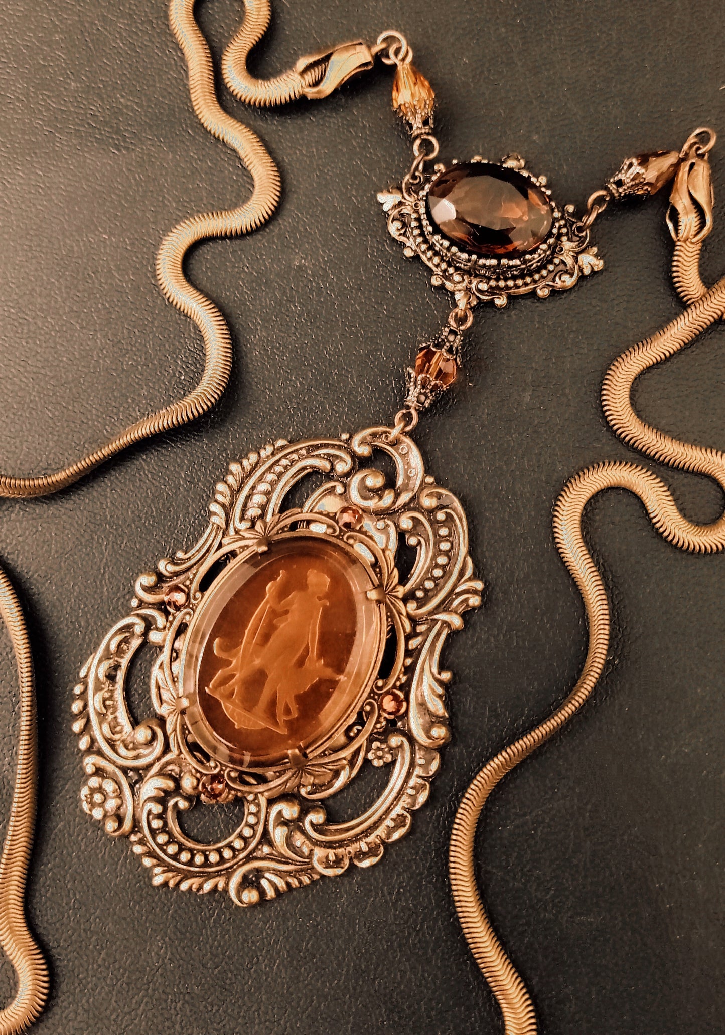 An ornate vintage necklace with a large amber cameo in an intricate brass filigree setting, connected by a smaller amber crystal medallion to gold snake chain.