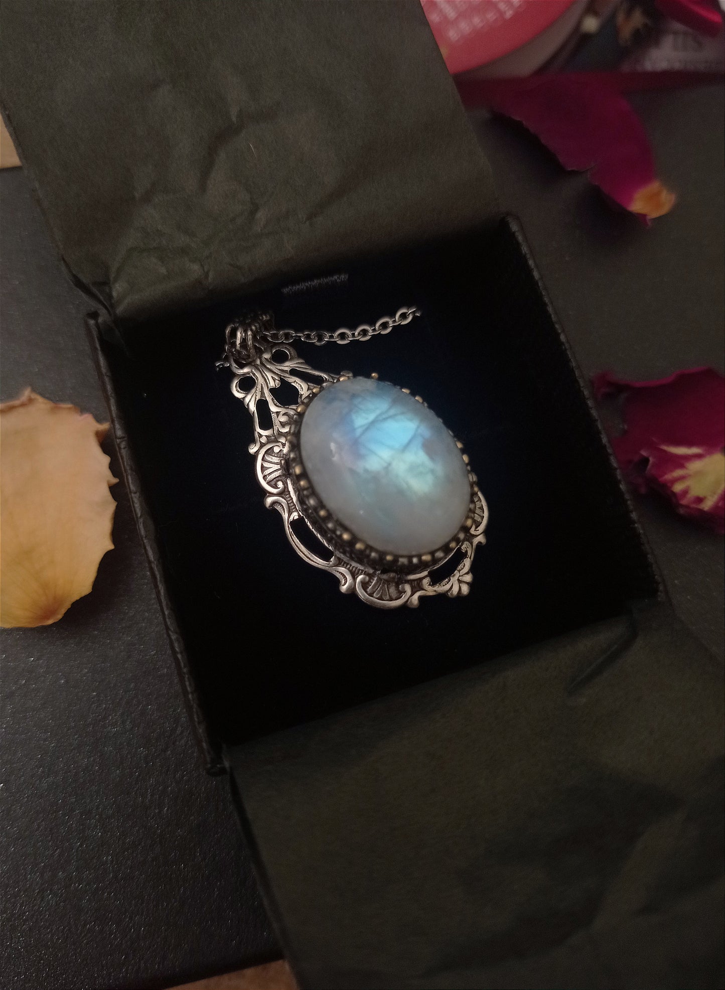 An elegant silver pendant necklace featuring a luminous real rainbow moonstone, set in an intricate vintage-style frame, displayed inside a black jewelry box, evoking timeless beauty and glamour.