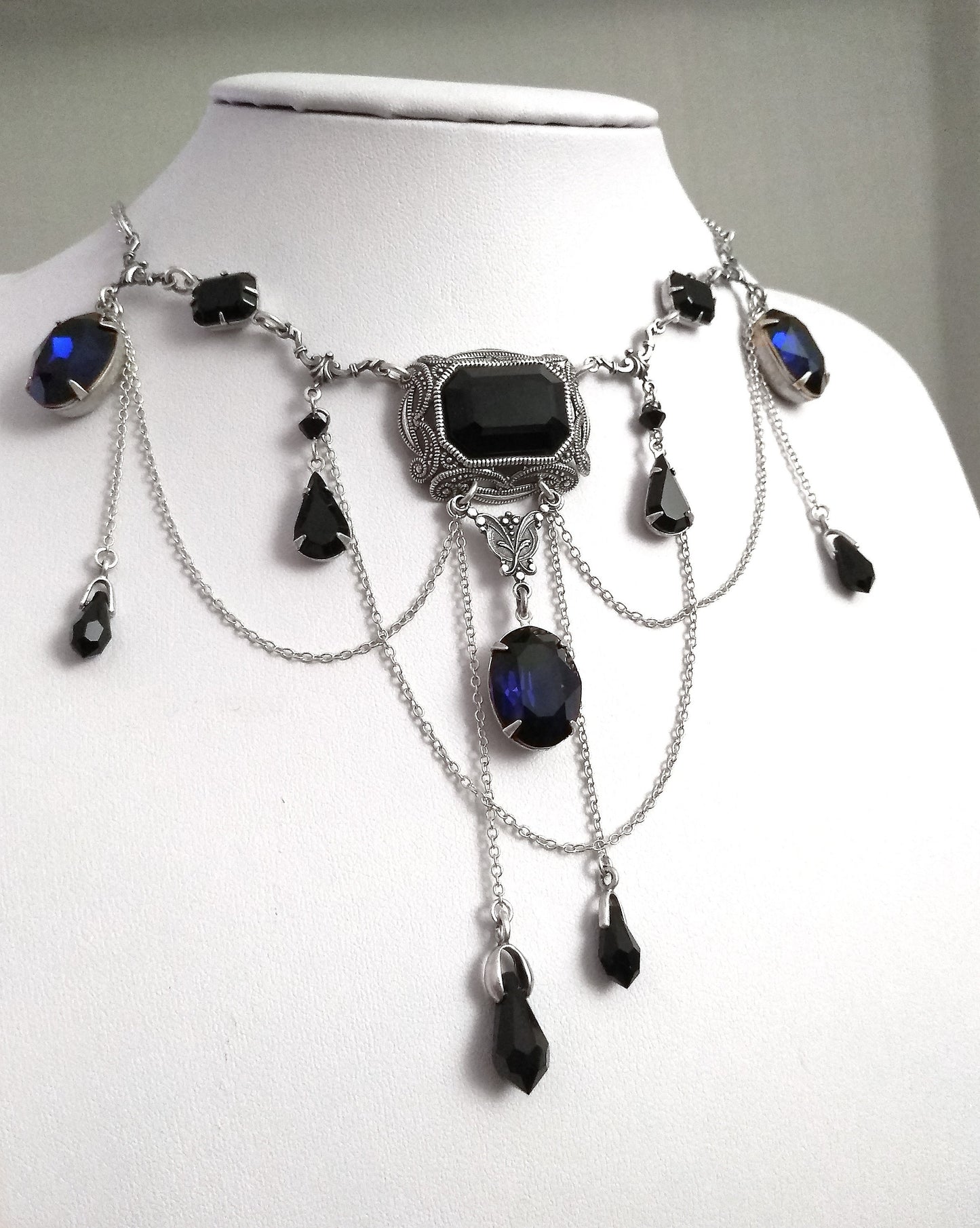 Gothic necklace with black and indigo Swarovski crystals, silver filigree centerpiece, and draping chains on bust display. Elegant gothic jewelry for women.