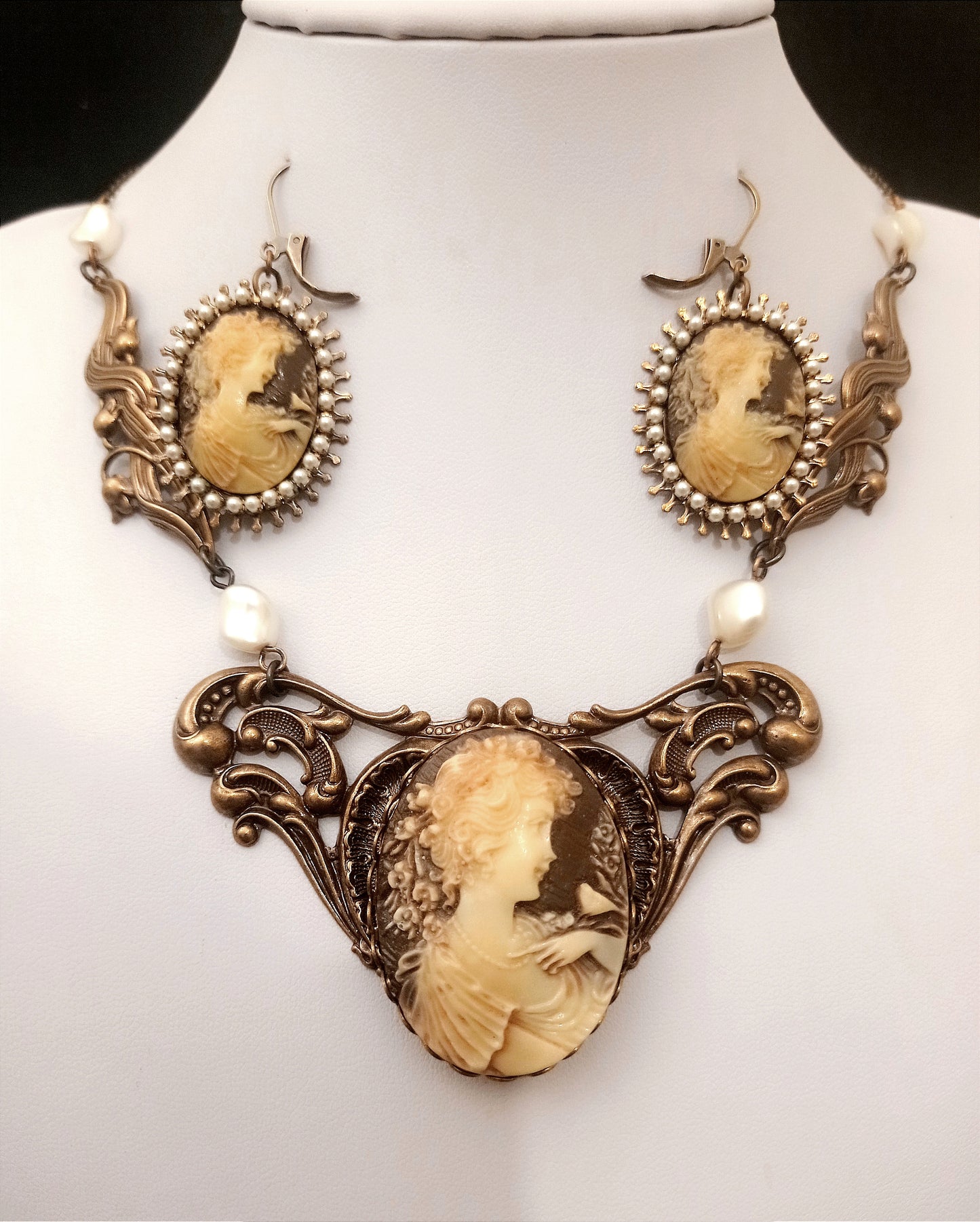 A Victorian style brass jewelry set features a cameo necklace and matching earrings. Each piece has a cream on brown portrait of a woman in profile, framed with ornate scrollwork and pearl accents