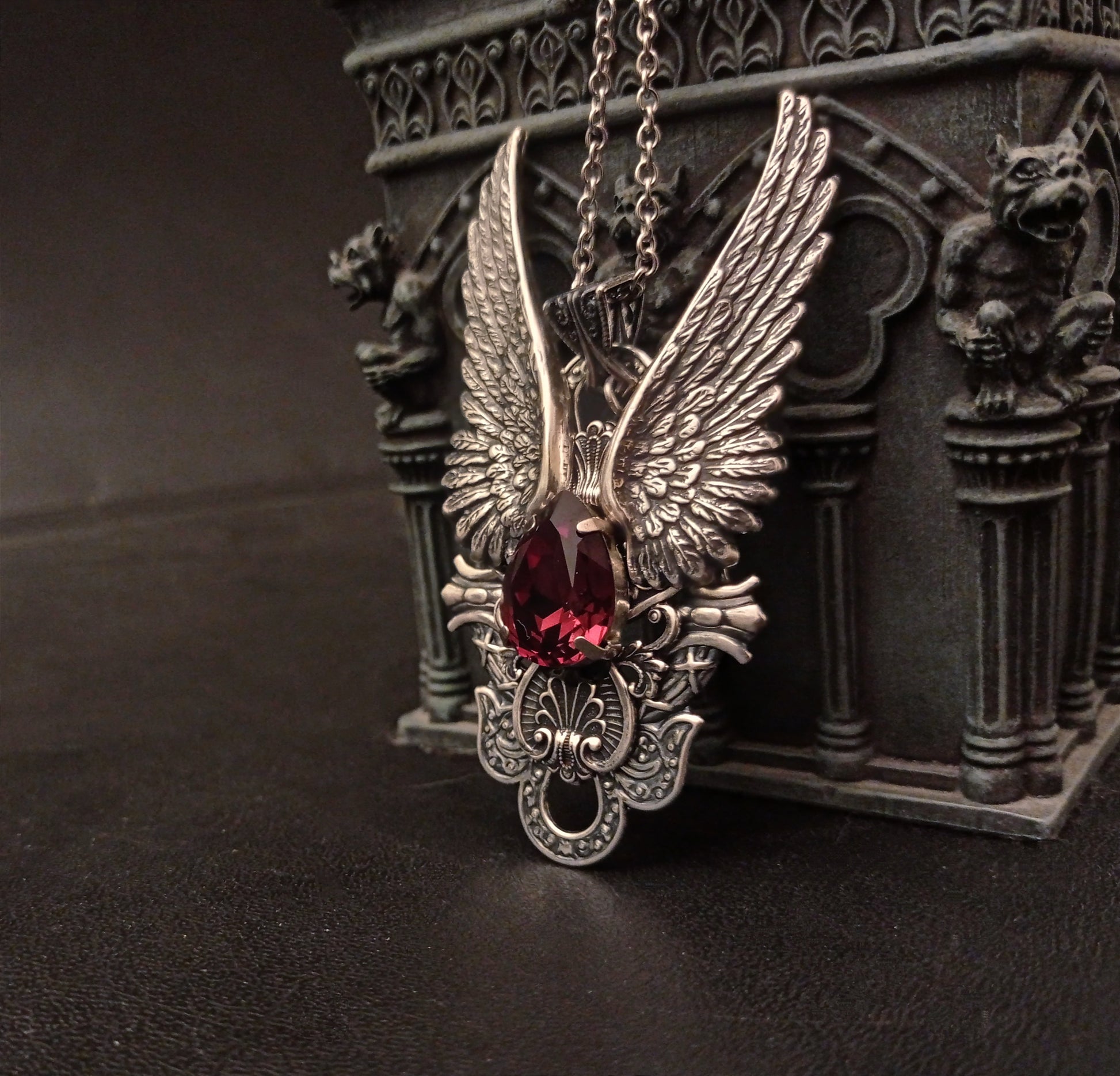 A large silver Gothic style pendant necklace featuring intricate wings and a large burgundy red gem.