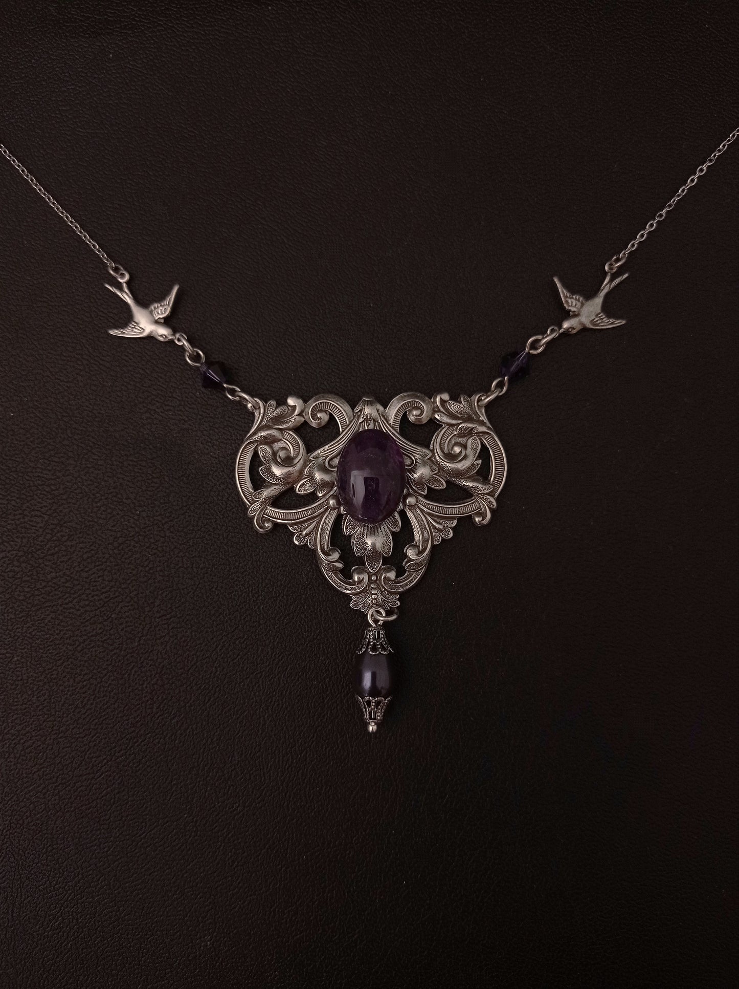 A Victorian style silver necklace with intricate floral patterns and two birds in flight, featuring an oval purple amethyst cabochon and smaller purple crystals and pearls.