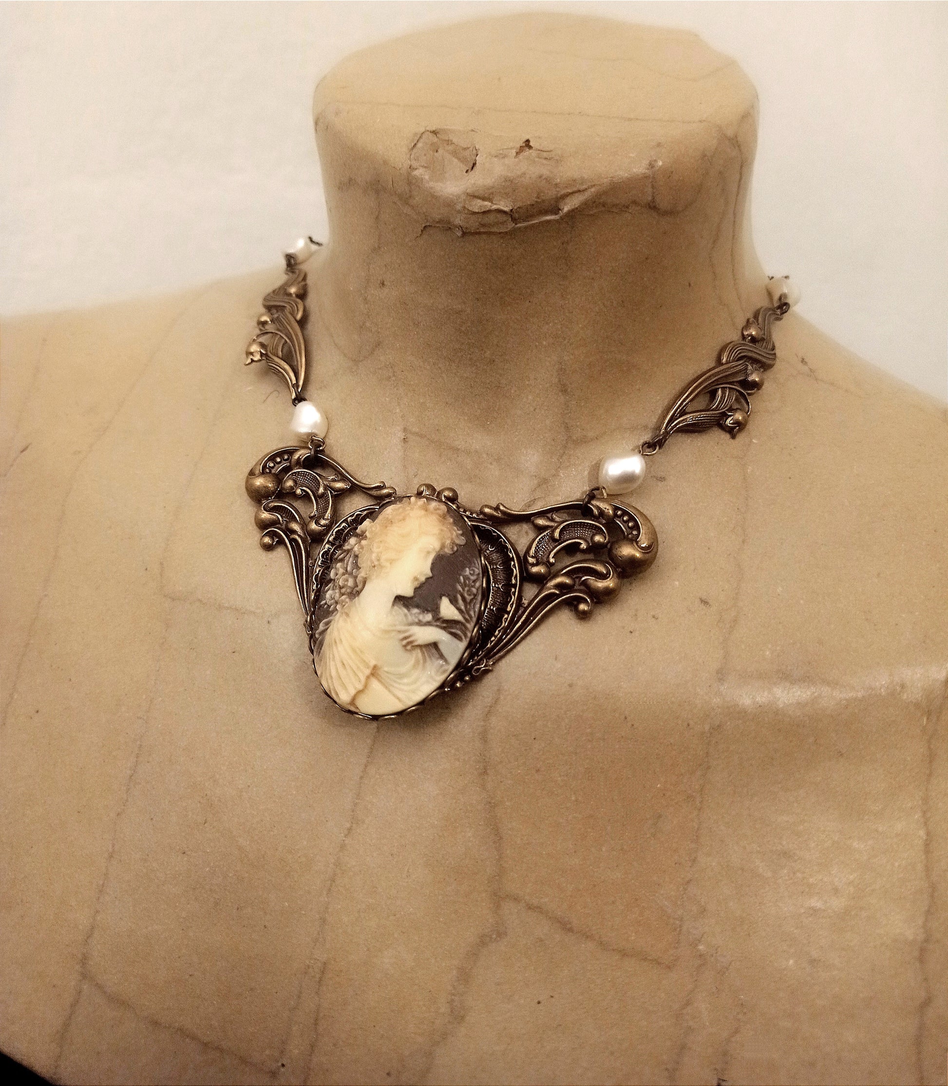 A Victorian style necklace featuring a prominent brass plaque with intricate patterns that hosts a brown cameo and is framed with pearls.