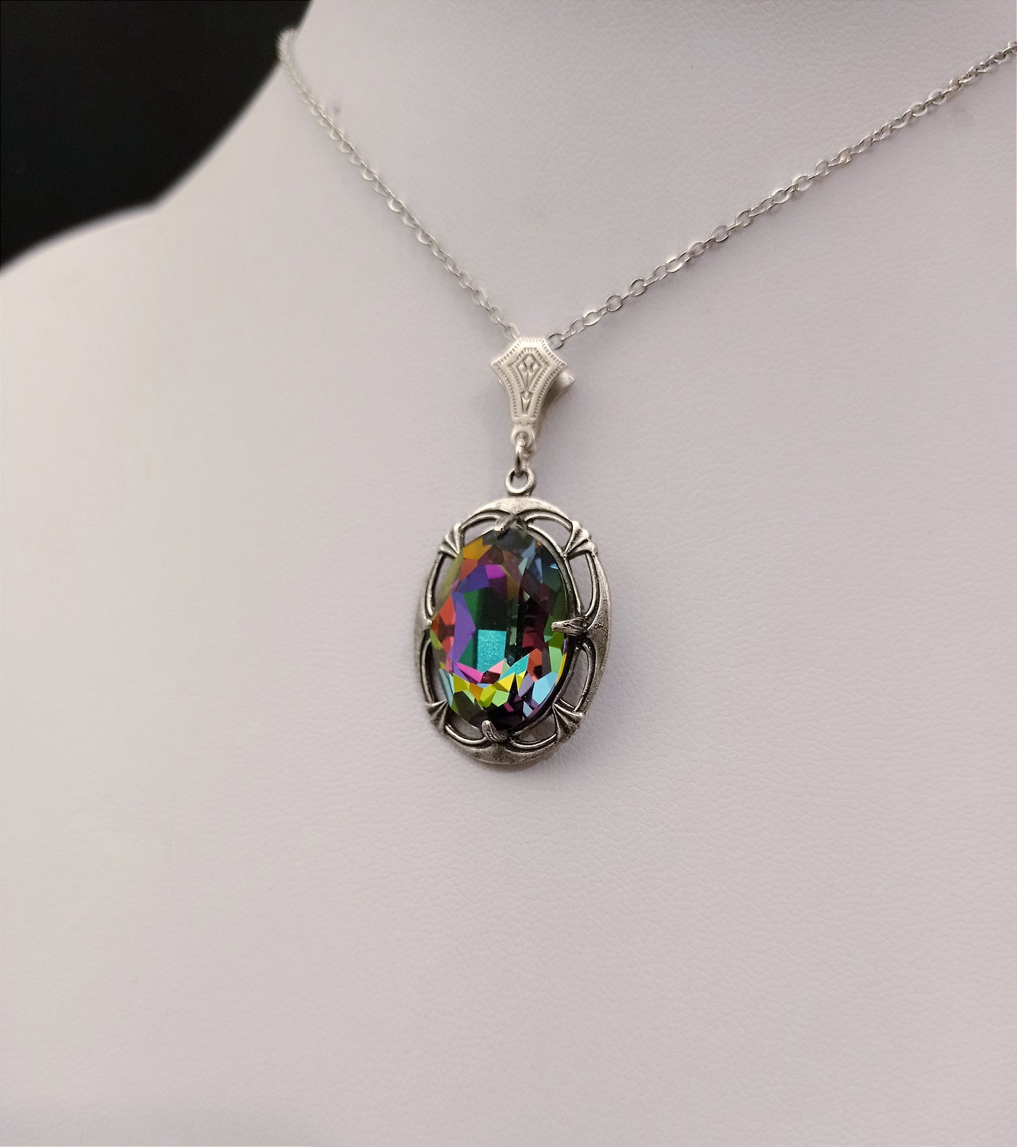 A colorful, iridescent oval crystal stone set in an ornate silver framework, hanging from a delicate chain.