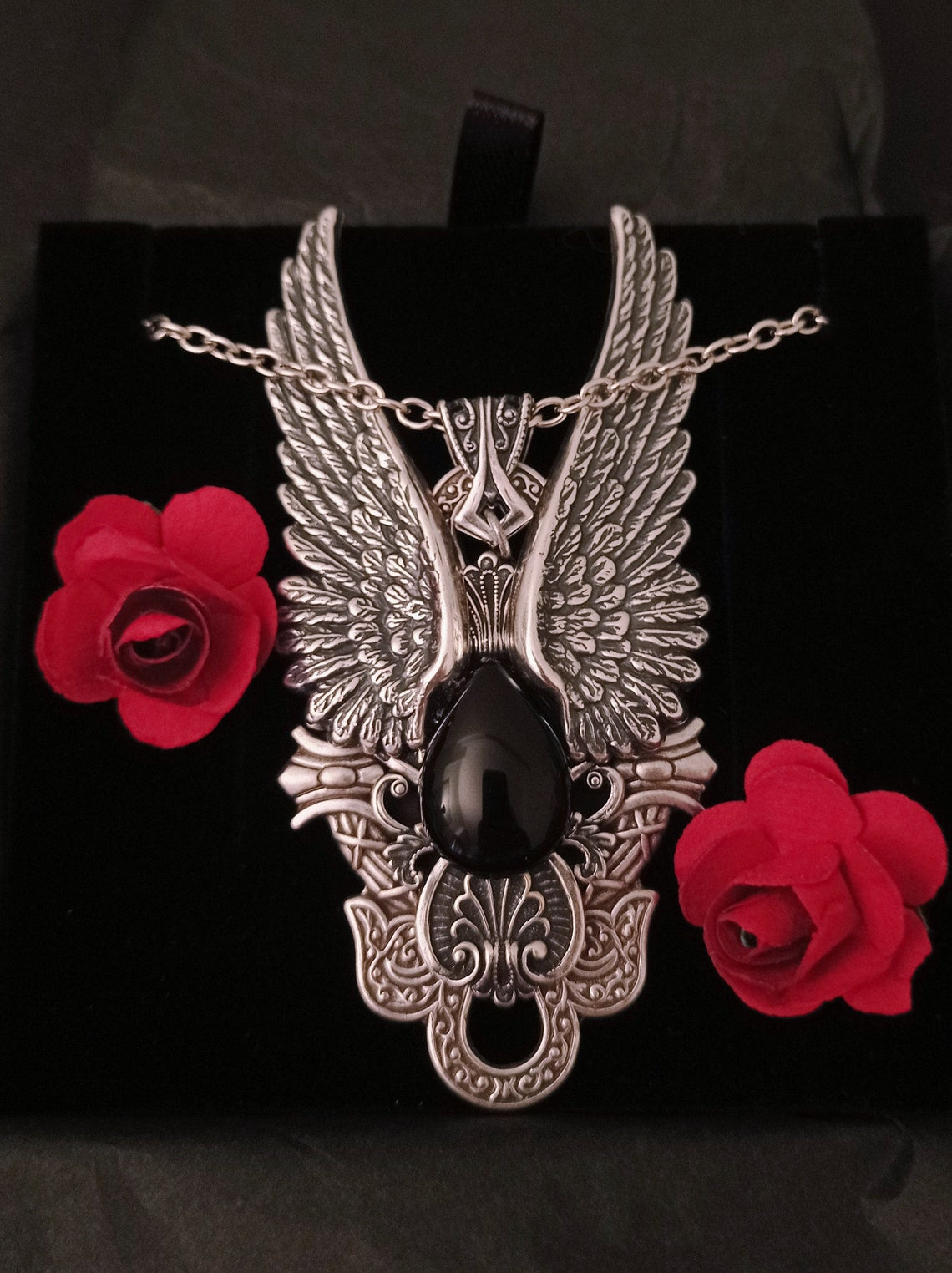 A Gothic pendant with ornate silver wings flanking a large, teardrop-shaped onyx cabochon. The piece features intricate decorative patterns and hangs from a matching silver chain.