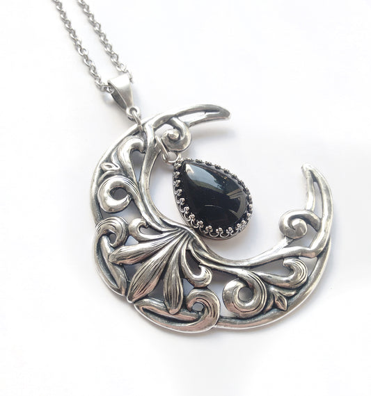 A silver-colored necklace featuring a detailed moon-shaped pendant with a black onyx centerpiece, surrounded by intricate celtic-like patterns. The pendant is attached to a chain.